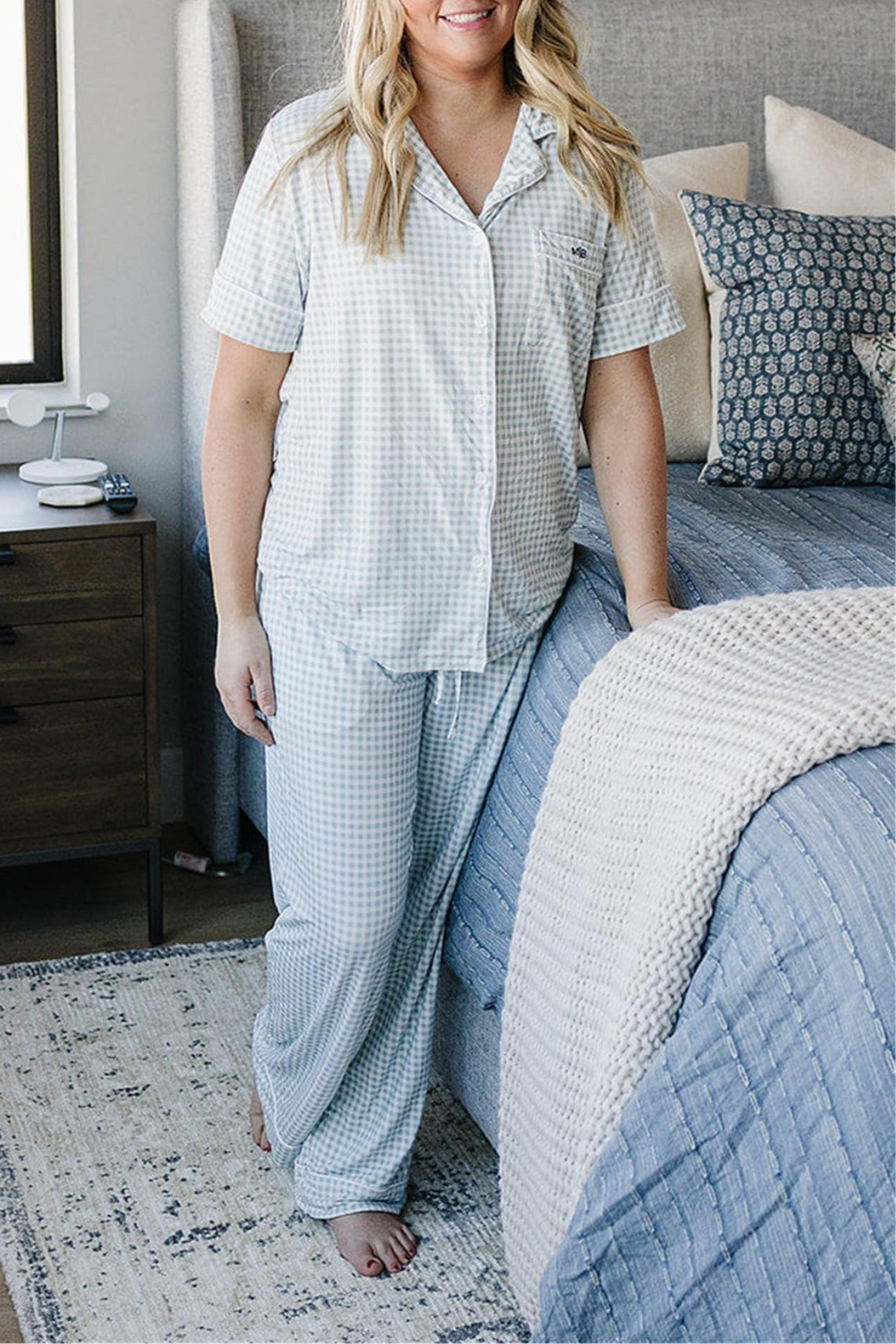 A woman in light blue gingham pajamas stands by a bed with blue and white bedding.
