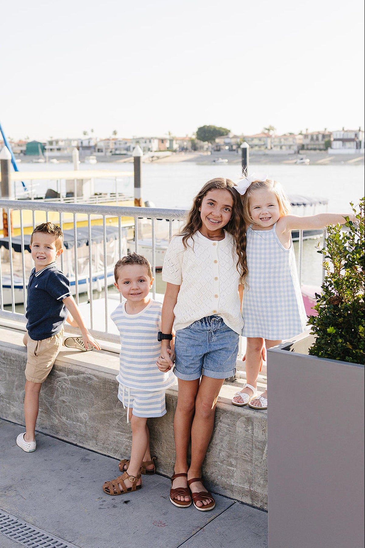 Four children stand by a waterfront, wearing summer outfits in various colors, smiling and enjoying the sunny day.