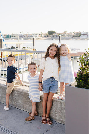 Four children stand by a waterfront, wearing summer outfits in various colors, smiling and enjoying the sunny day.
