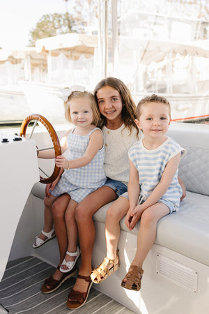 Three children sit on a boat, wearing stylish summer outfits in light blue and white patterns, enjoying a sunny day.