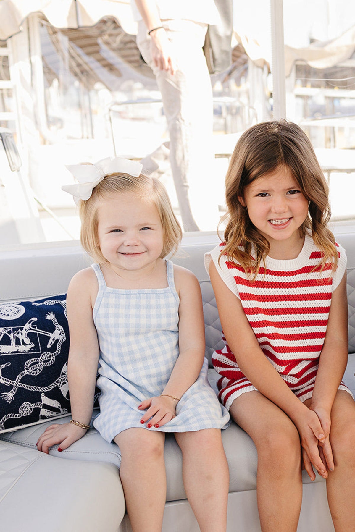 Two young girls sit on a boat, one in a light blue gingham dress and the other in a red.