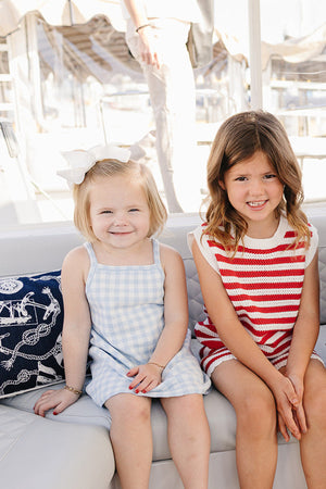 Two young girls sit on a boat, one in a light blue gingham dress and the other in a red.