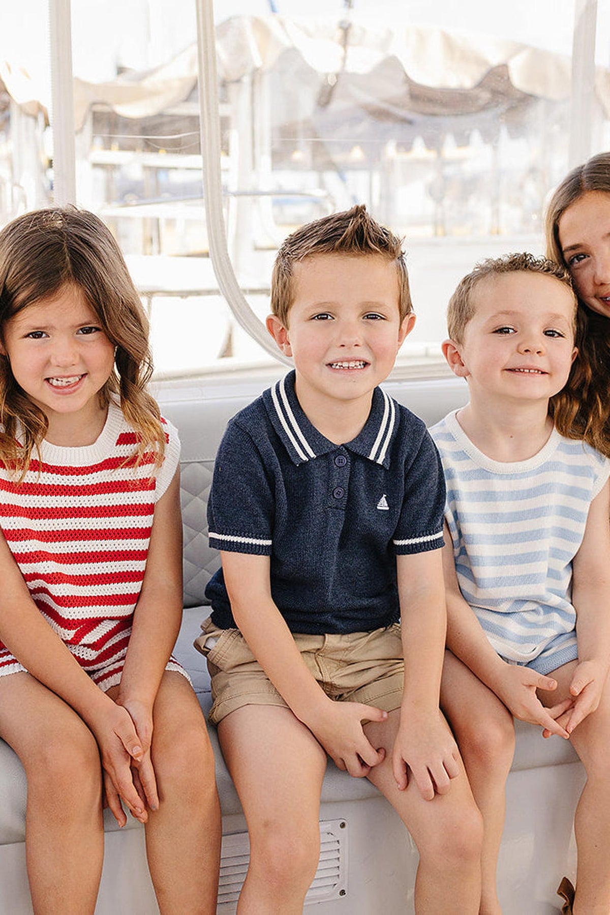 Four children sit together on a boat, wearing colorful summer outfits, smiling against a sunny, coastal backdrop.