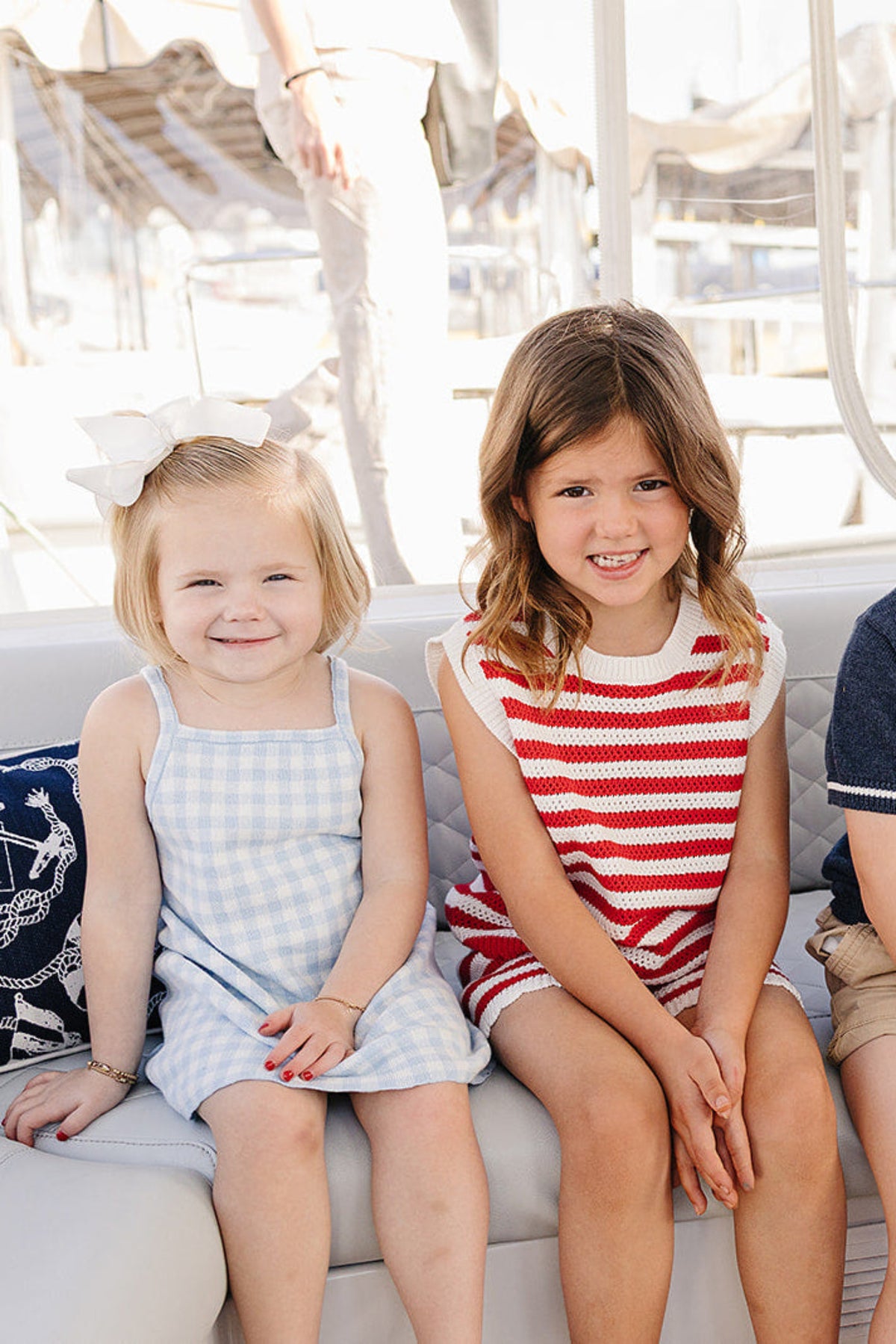 Two young girls sit side by side on a boat, one wearing a blue and white gingham dress and the other in a red and white striped knit tank set.