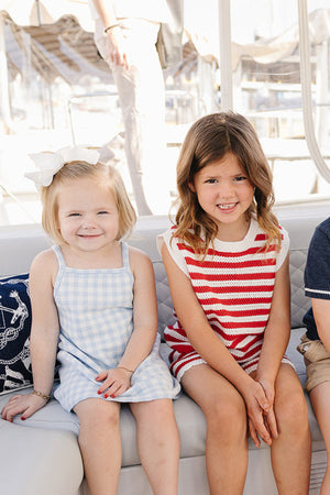 Two young girls sit side by side on a boat, one wearing a blue and white gingham dress and the other in a red and white striped knit tank set.