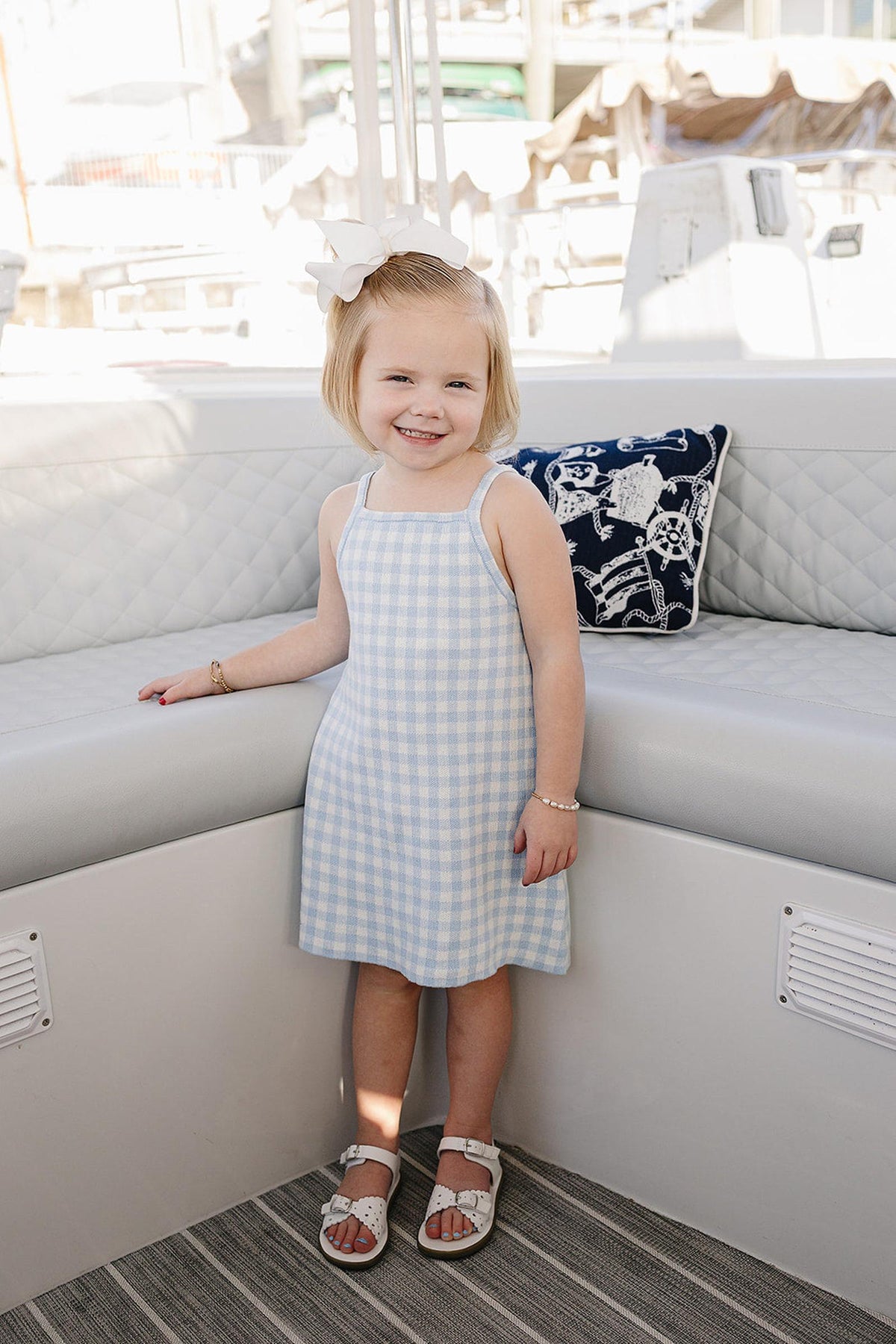 A young girl smiles while wearing a light blue gingham dress, paired with white sandals, in a boat setting.
