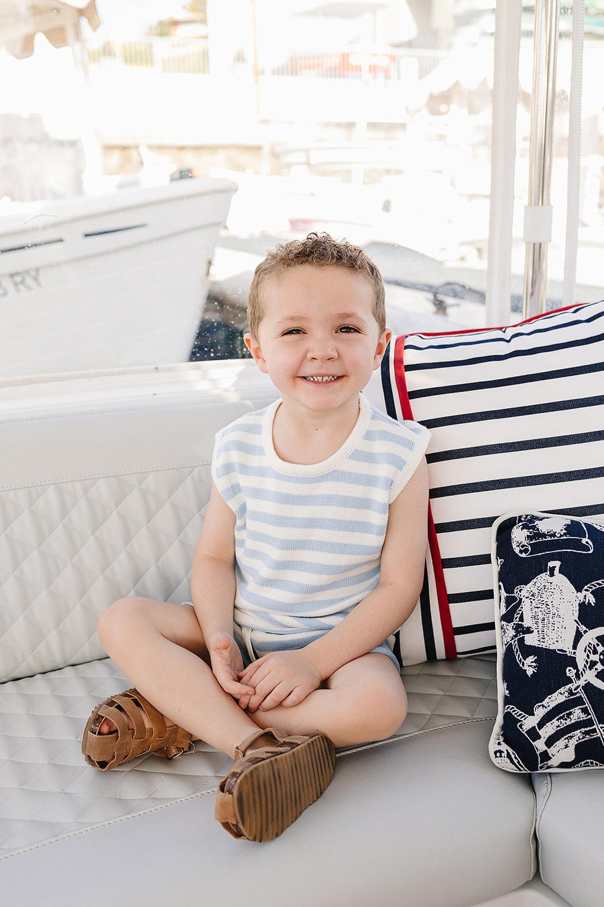 A smiling child sits on a cushioned seat, wearing a blue and white striped tank top and brown sandals.