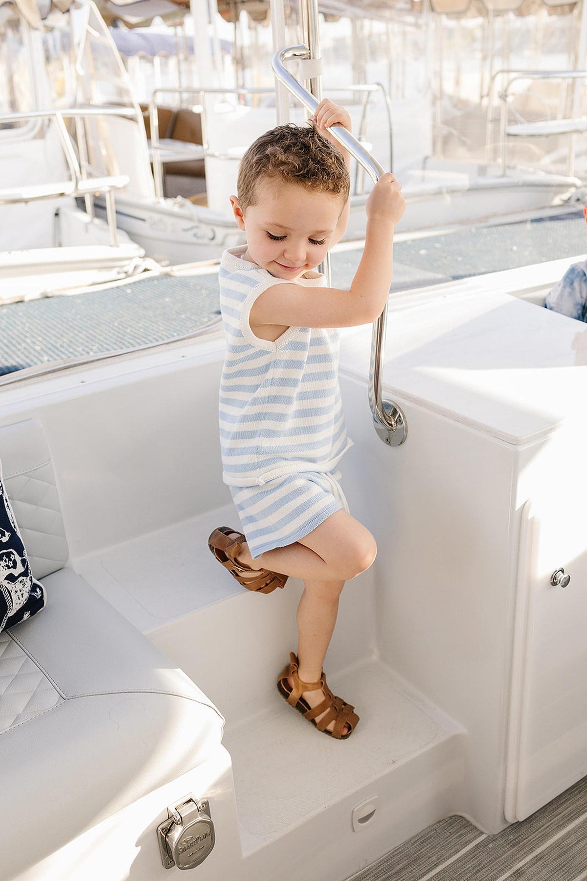 A young child wearing a blue and white striped tank set stands on a boat, holding onto a metal railing.