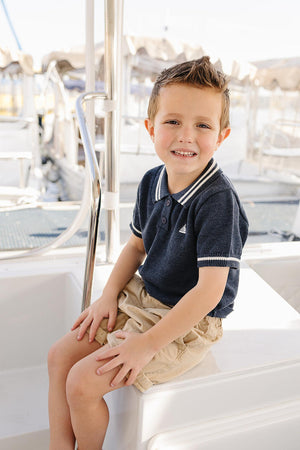A young boy sits on a boat, wearing a navy collared knit shirt with white trim and beige shorts, smiling.