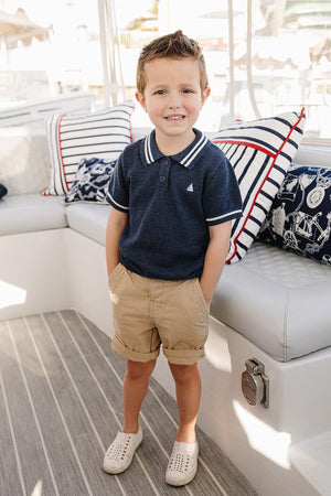 A young boy stands on a boat, wearing a navy collared shirt, khaki shorts, and white slip-on shoes, smiling.