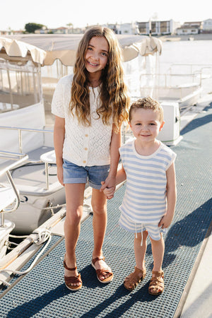 A girl in a white knit cardigan and denim shorts stands next to a boy in a striped tank top,.