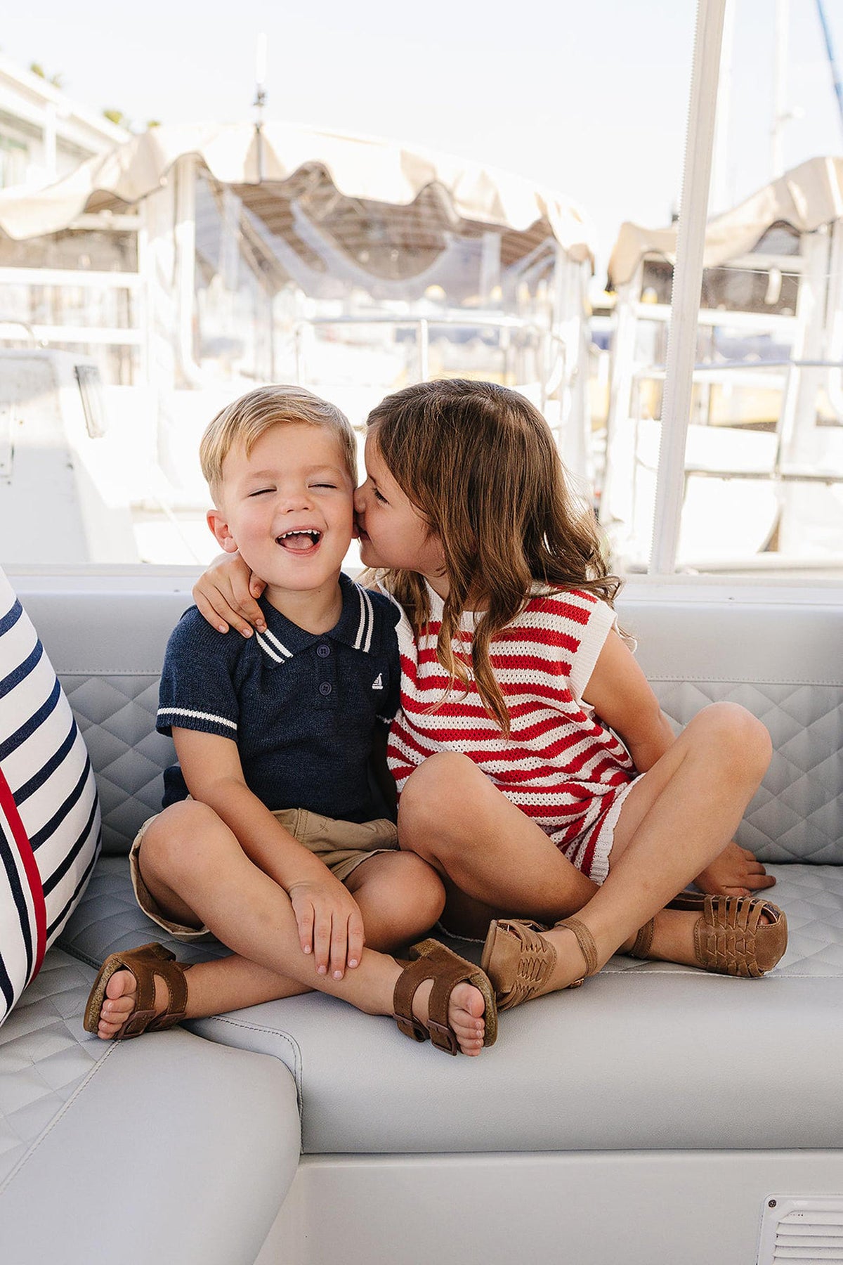 Two children sit together on a boat, with one girl playfully kissing the boy on the cheek. They wear casual summer outfits, showcasing a relaxed and joyful moment.