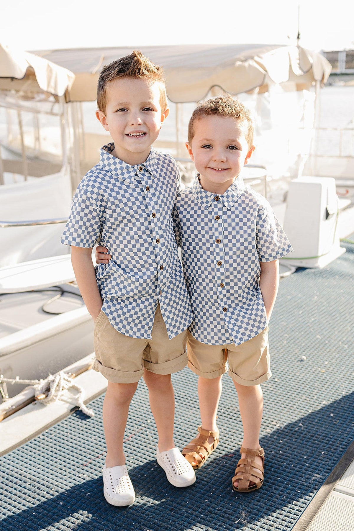 Two young boys stand side by side on a boat dock, wearing light blue checkered button-up shirts and beige shorts, smiling brightly against a backdrop of boats and water.