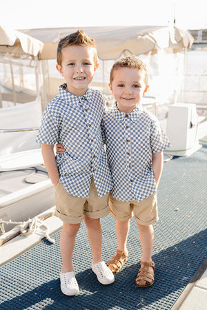 Two young boys stand side by side on a boat dock, wearing light blue checkered button-up shirts and beige shorts, smiling brightly against a backdrop of boats and water.