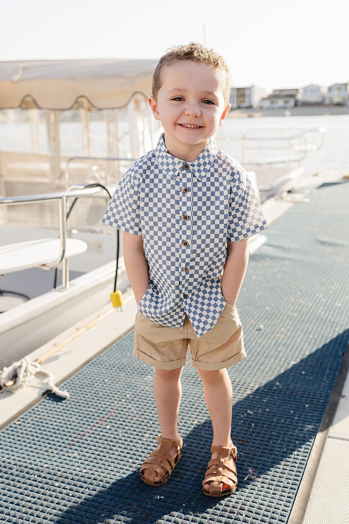 A young boy stands on a dock, wearing a light blue checkered button-up shirt and beige shorts, smiling confidently.