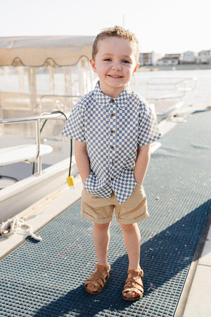 A young boy stands on a dock, wearing a light blue checkered button-up shirt and beige shorts, smiling confidently.