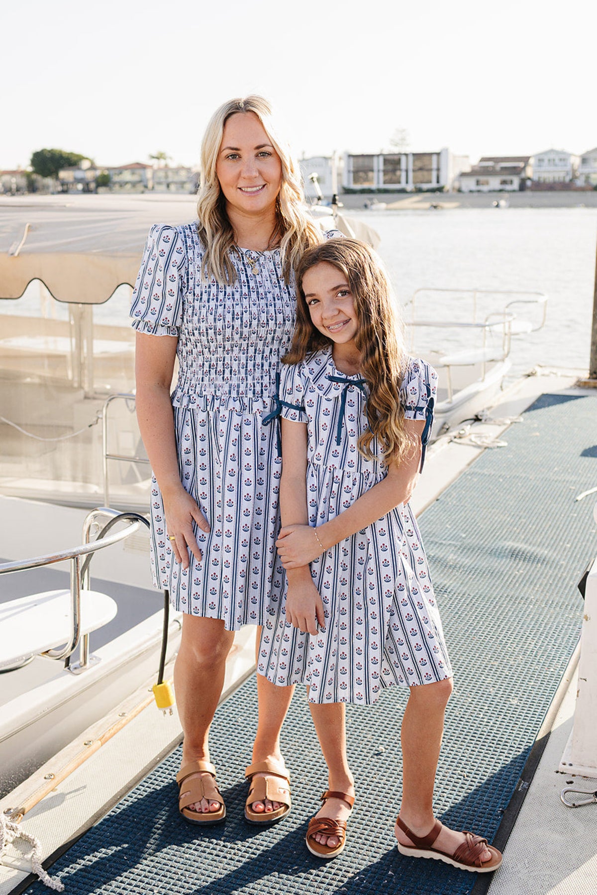 A woman and a girl stand on a dock, both wearing matching striped dresses with floral patterns. The background features a calm waterfront with boats, creating a serene setting.