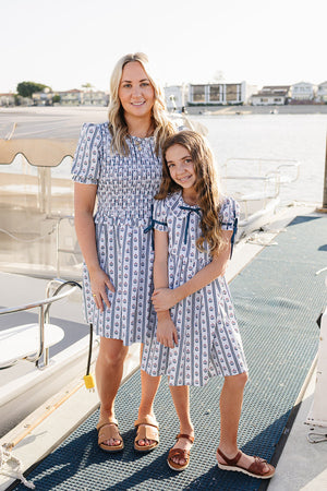 A woman and a girl stand on a dock, both wearing matching striped dresses with floral patterns. The background features a calm waterfront with boats, creating a serene setting.