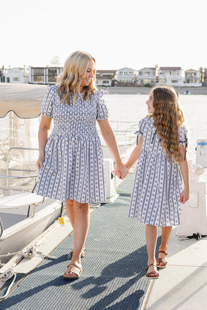 A mother and daughter walk hand-in-hand on a boat dock, both wearing matching blue and white block print dresses.