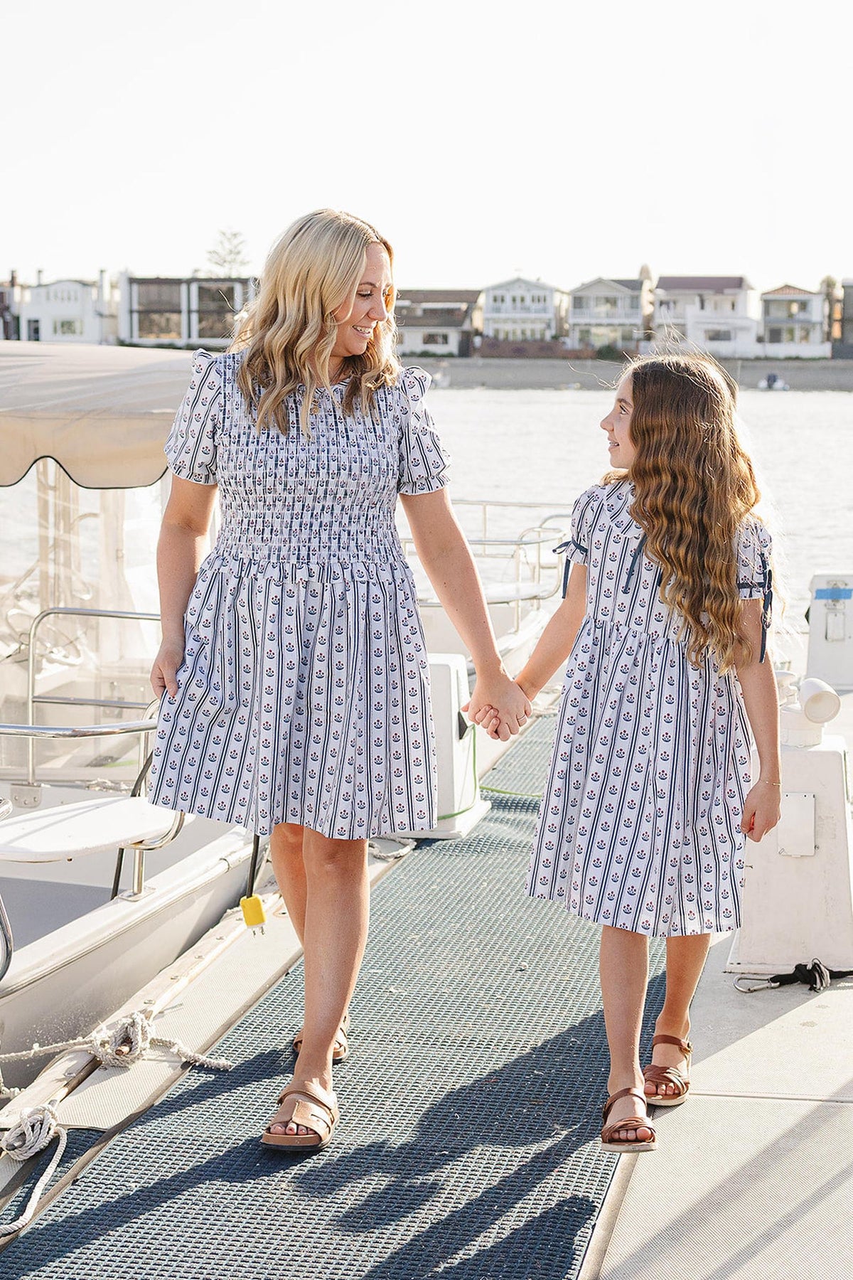 A woman and a girl walk hand-in-hand on a dock, both wearing matching blue and white striped floral dresses. The scene features a serene waterfront with homes in the background.