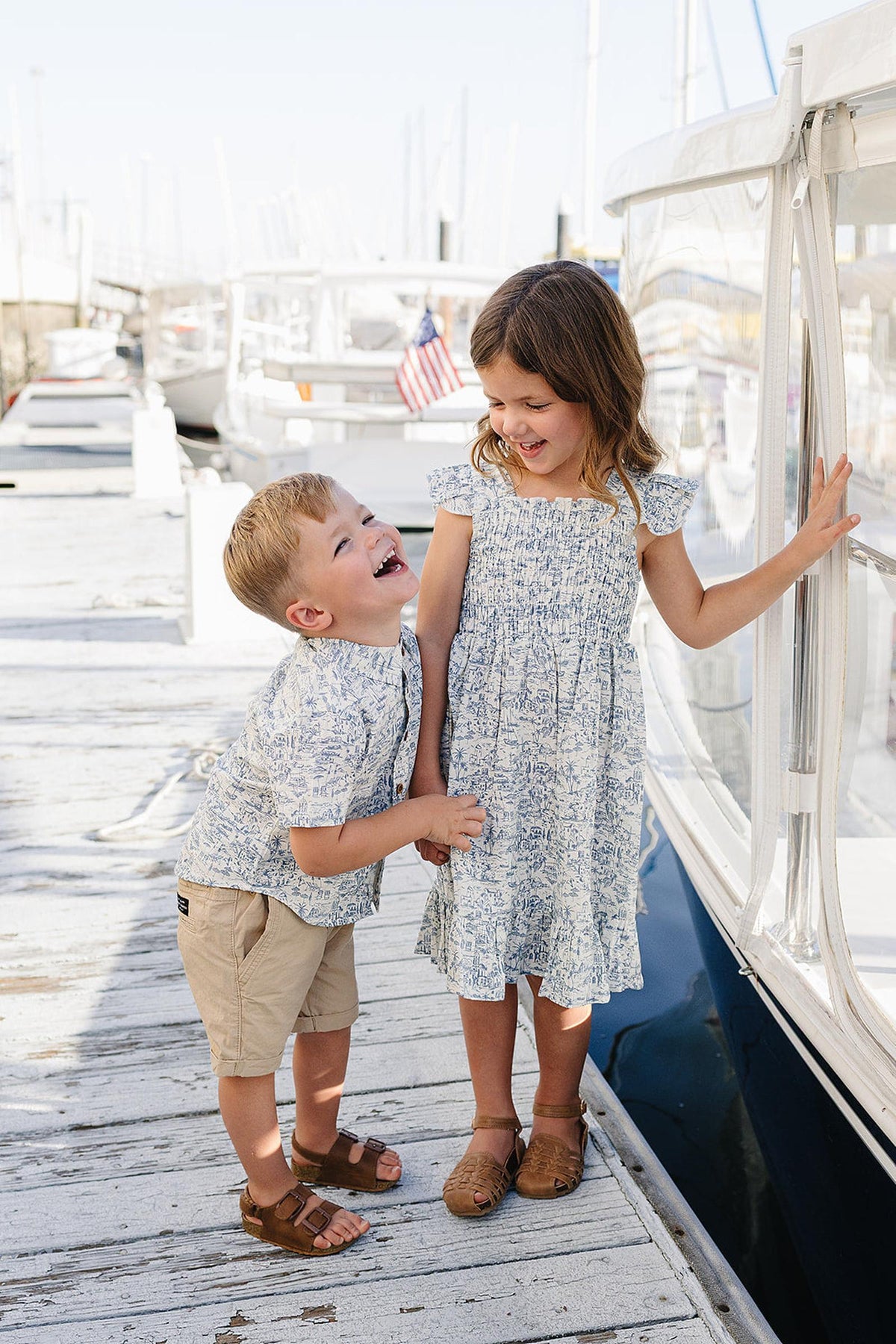 Two children play on a dock by a marina, with the boy wearing a light blue patterned button-up shirt and the girl in a matching dress.