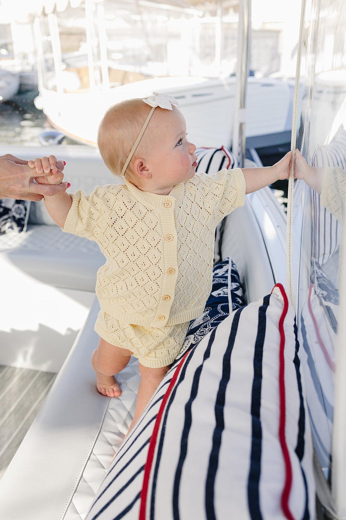 A baby girl in a yellow knit cardigan and bloomer set reaches toward a reflective surface, sitting on a boat with striped cushions.