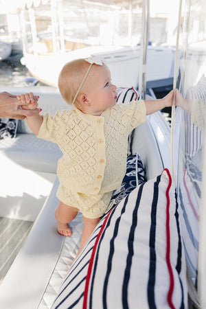 A baby girl in a yellow knit cardigan and bloomer set reaches toward a reflective surface, sitting on a boat with striped cushions.