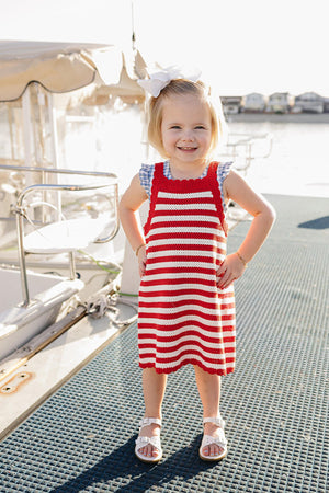 A smiling young girl stands on a dock, wearing a red and white striped knit dress with scalloped edges.