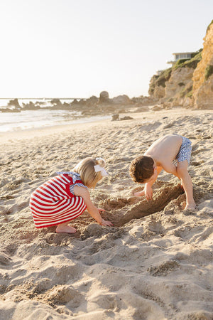 Two children play on a sandy beach, digging in the sand. One girl wears a red and white striped dress, while a boy in blue shorts focuses on their sandcastle.