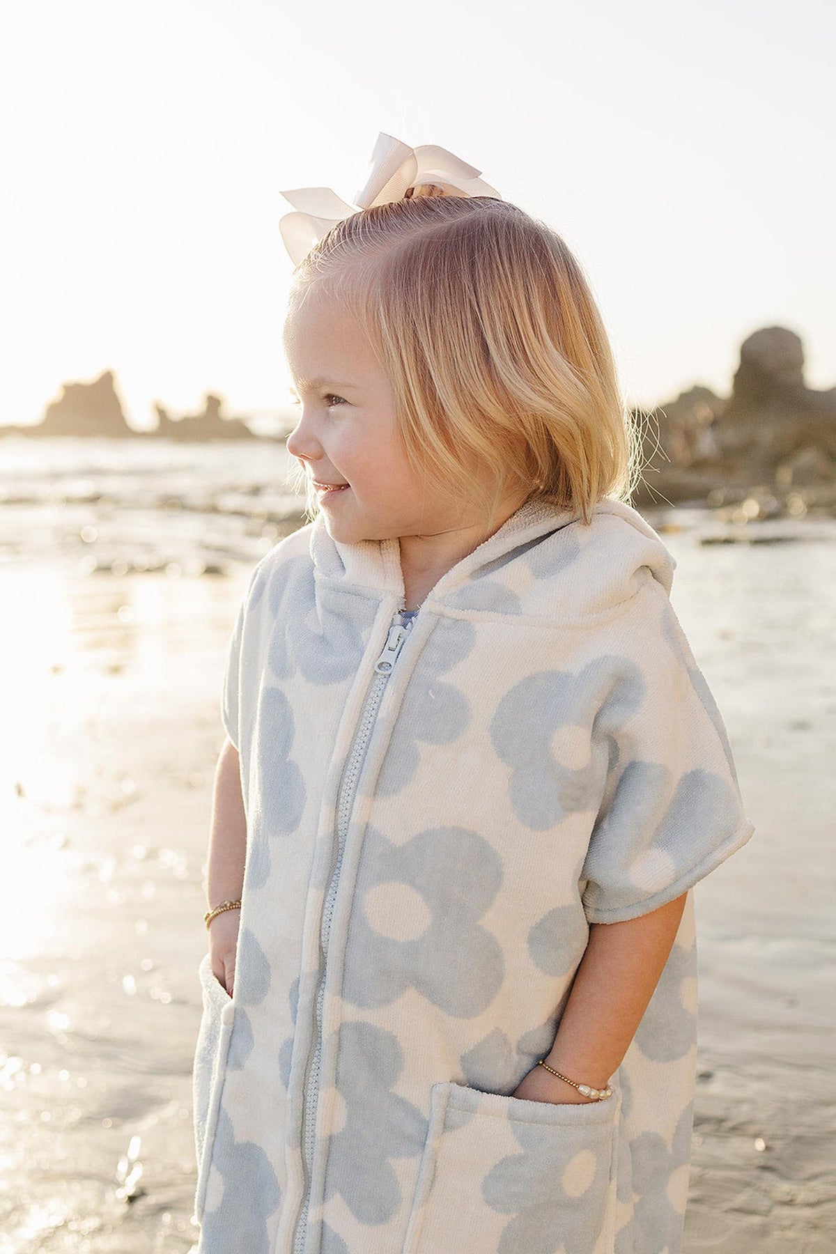 A young child wearing a light blue and white floral zip-up beach poncho stands by the water, smiling. The poncho features a hood and pockets, made from soft, organic cotton.