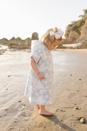 A child wearing a light blue and white floral zip-up poncho stands barefoot on a sandy beach, with soft waves in the background and a sunny coastline.