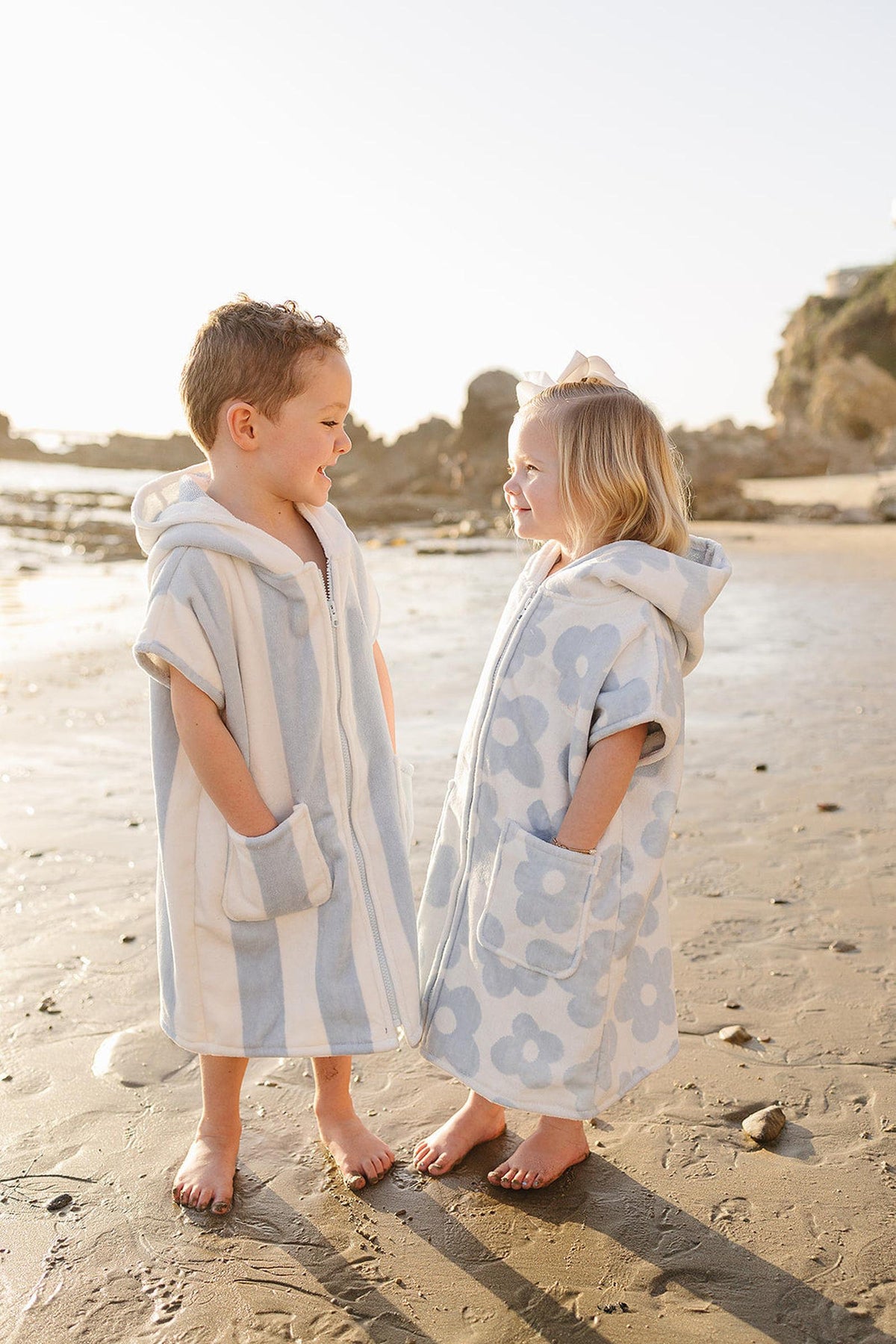 Two children stand on a sandy beach, wearing light blue and white zip-up beach ponchos made from organic cotton. The sun sets in the background, creating a warm, inviting atmosphere.