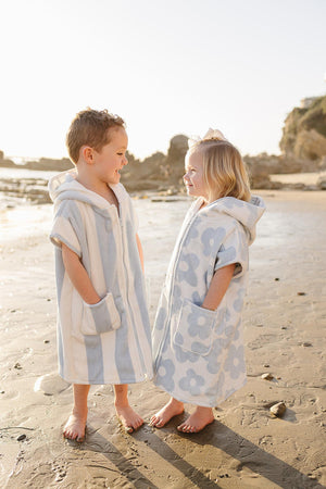 Two children stand on a sandy beach, wearing light blue and white zip-up beach ponchos made from organic cotton. The sun sets in the background, creating a warm, inviting atmosphere.