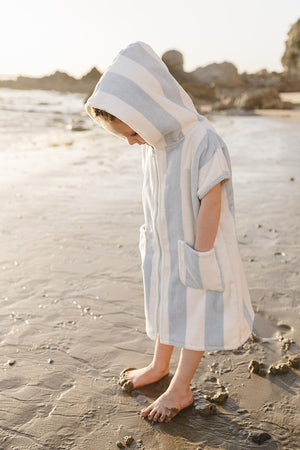 A child stands on wet sand by the beach, wearing a light blue and white striped zip-up poncho with a hood.