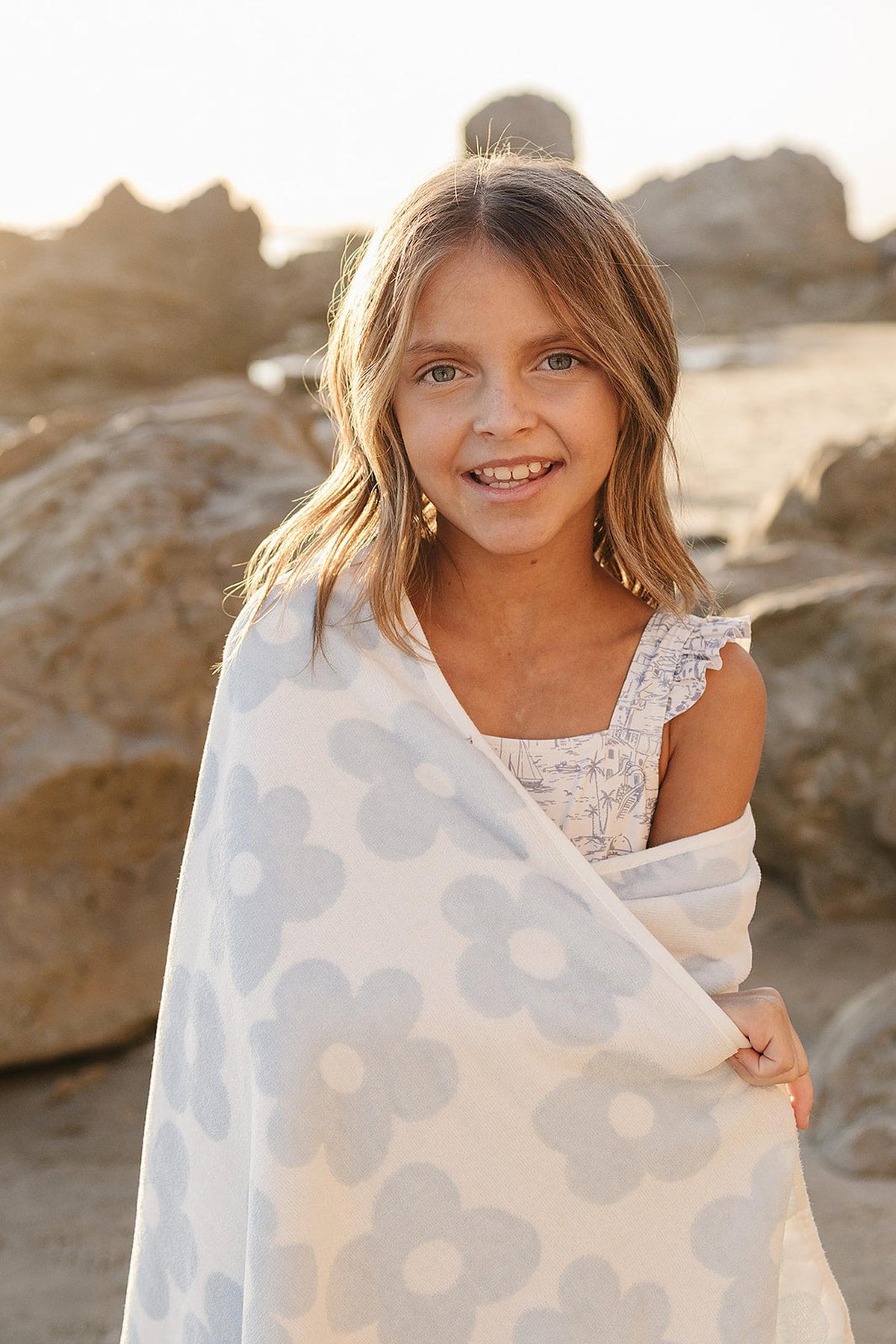 A young girl smiles while wrapped in a light blue and white floral towel, standing on a sandy beach with rocky formations in the background during golden hour.