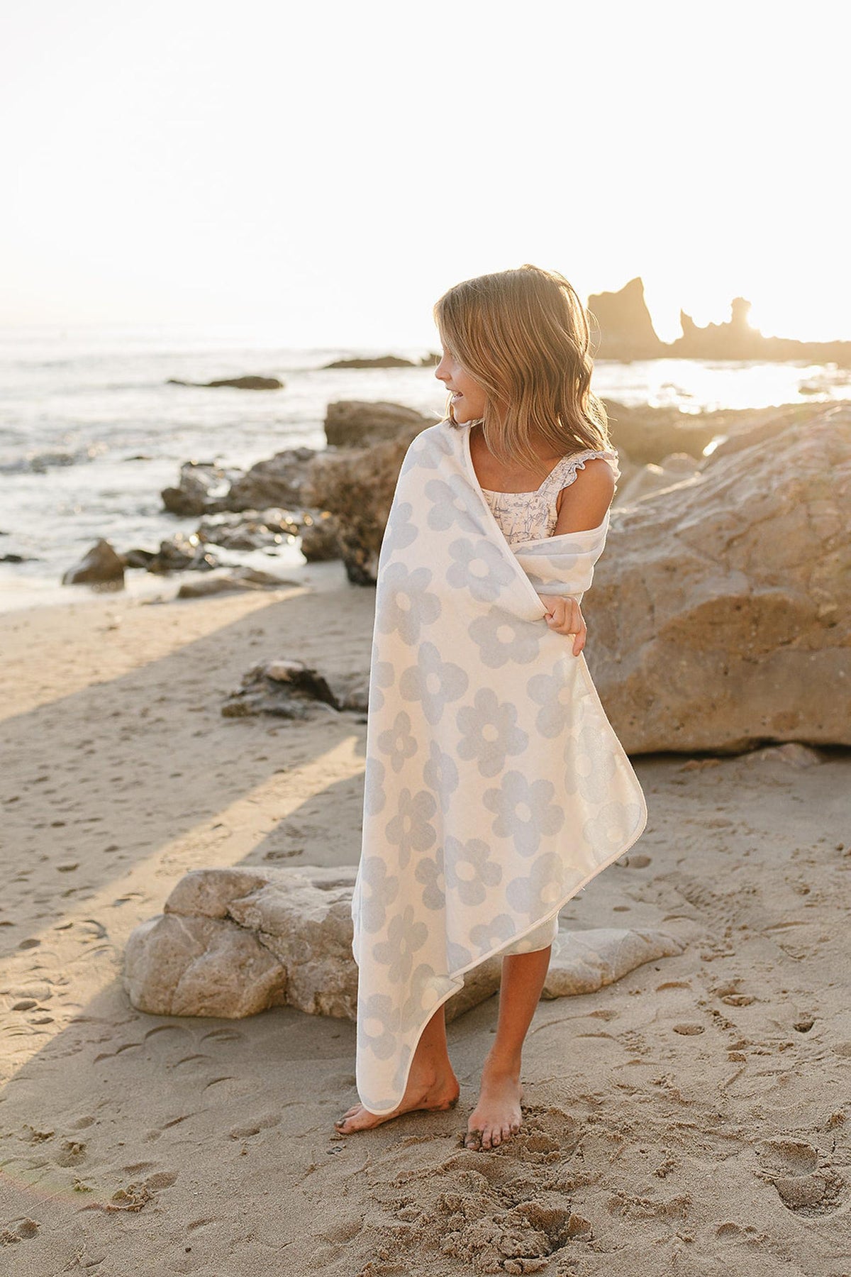 A young girl stands on a sandy beach, wrapped in a light blue and white floral organic cotton beach towel, with sunlit waves and rocky formations in the background.