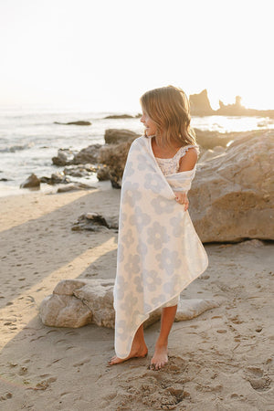 A young girl stands on a sandy beach, wrapped in a light blue and white floral organic cotton beach towel, with sunlit waves and rocky formations in the background.