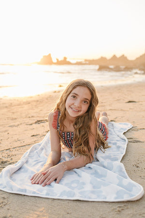 A young girl lies on a light blue beach towel with a floral pattern, enjoying the sandy beach during sunset. The towels organic cotton material is soft and eco-friendly.