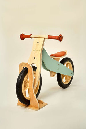A wooden balance bike stand displays a light wood finish with smooth edges, designed to hold a colorful balance bike featuring a mint green frame and orange seat, perfect for.