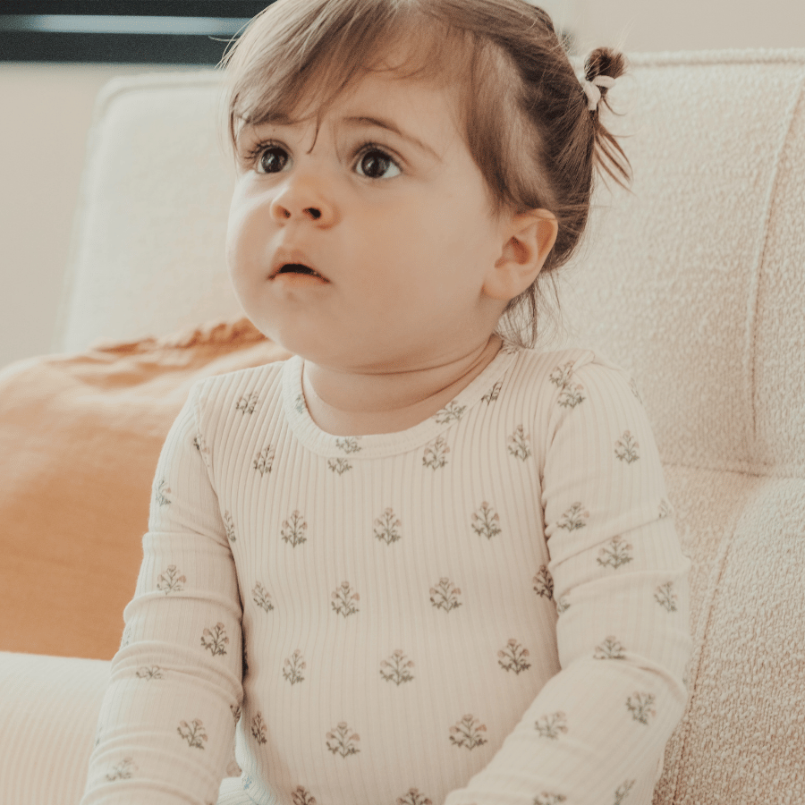 A baby sits on a light-colored sofa, wearing a soft, long-sleeved romper with a delicate floral print. The romper is made from cotton, providing comfort for infants.