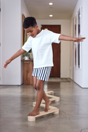 A young boy balances on a wooden balance beam in a modern hallway. He wears a white t-shirt and striped shorts, showcasing active play and coordination skills.