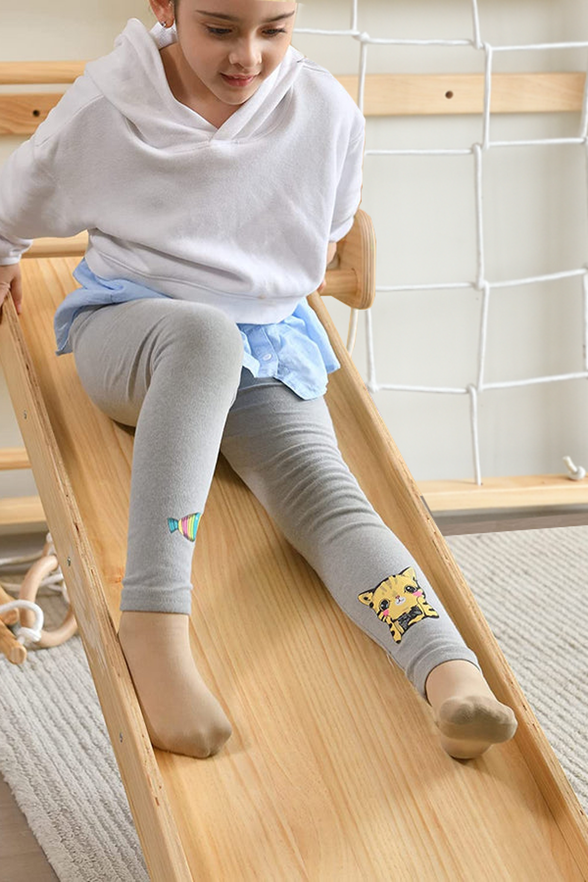 A child wearing a white hoodie and gray leggings slides down a wooden ramp with playful designs.