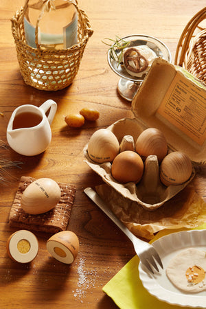 A wooden egg playset is displayed on a rustic wooden table, featuring various wooden eggs in an egg carton, alongside a small pitcher and decorative items, promoting imaginative play.