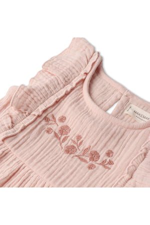 Close-up of a blush-colored organic cotton dress featuring delicate floral embroidery and soft, tiered ruffles. The dress is designed for children, emphasizing a sustainable and charming aesthetic.