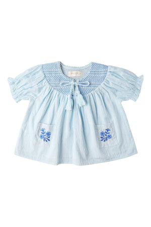 Light blue organic cotton smocked top featuring delicate embroidery on the pockets and a textured dobby weave. Designed for comfort, it is perfect for sunny day outings.