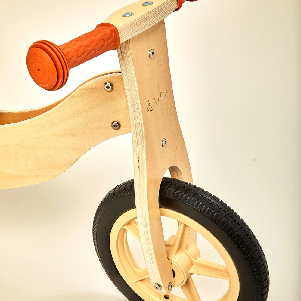 A close-up view of a wooden balance bike featuring a natural wood finish, orange rubber grips, and a sturdy wheel design.