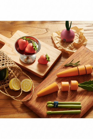 A wooden cutting board displays vibrant wooden strawberries, a playful carrot set, and a bowl with colorful produce. The eco-friendly play food encourages imaginative kitchen adventures for children.