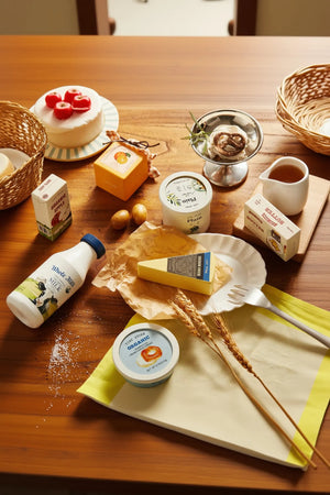 A wooden table is set with various dairy products, including milk bottles, yogurt containers, and cheese, alongside decorative items like pastries and eggs, creating an inviting and natural food scene.