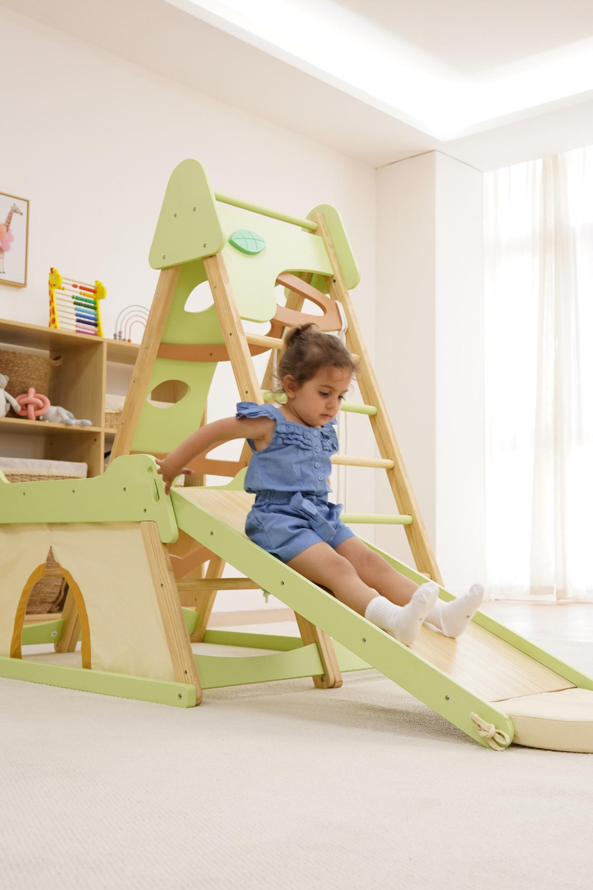 A toddler in a blue outfit slides down a wooden jungle gym with a green and natural finish.