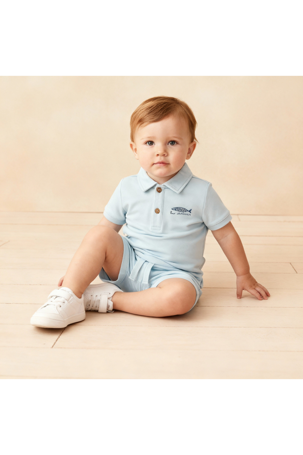 A young child sits on a light wooden surface, wearing a light blue organic polo shirt and matching shorts.